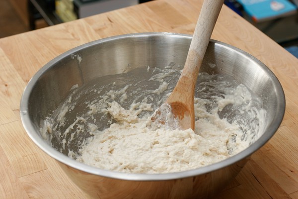 A metal mixing bowl with dough and a wooden spoon in it