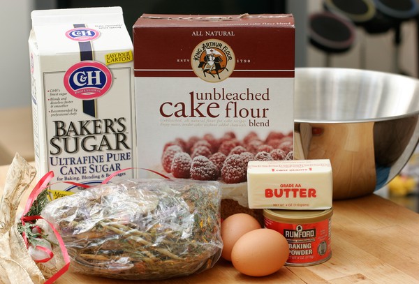 A box of ultra-fine baker's sugar, a box of unbleached cake flour, a metal mixing bowl, a bag of potpourri, two brown eggs, a stick of butter, and a tin of baking powder