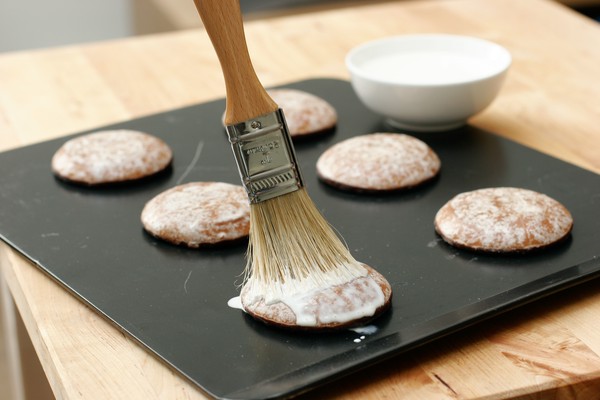 A pastry brush applying a white glaze to six cookies on a baking sheet; the bowl of glaze is in the background; the cookies now look perfect and have no obvious potpourri sticking out