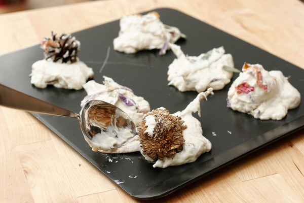 Six mounds of dough with large pieces of potpourri sticking out of them on a cookie sheet; a metal spoon is depositing the last mound