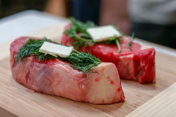 Two New York strip steaks topped with dill, tarragon, and a pat of butter on a wooden cutting board