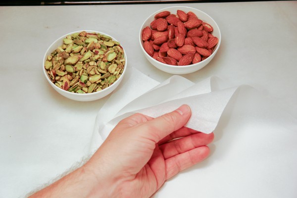 A human hand putting 3 sheets of parchment on a counter, next to a small white bowl of chopped pistachios and a small white bowl of whole almonds