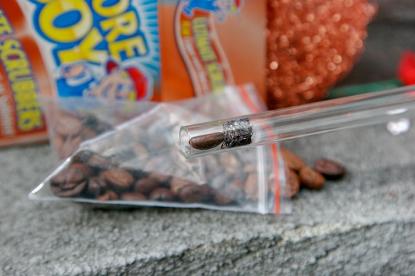 A glass tube with a piece of scouring pad and a coffee bean inside it; a bag of coffee beans is in the background along with an open box of scouring pads