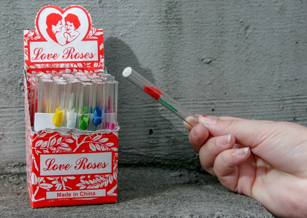 A human hand holding a glass tube with a red, plastic rose inside; an open box of multicolored love roses is in the background
