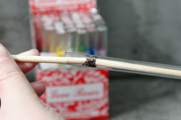 A human hand using a chopstick to pack a piece of burnt scouring pad into a glass tube; an open box of multicolored love roses is in the background