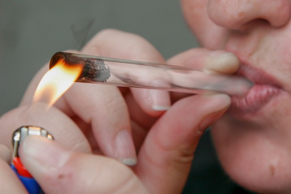 A human holding and inhaling from a glass tube with a coffee bean and scouring pad piece inside of it; one hand holds a lighter such that the flame is hitting the end of the tube with the coffee bean; smoke travels down the tube into the human's mouth