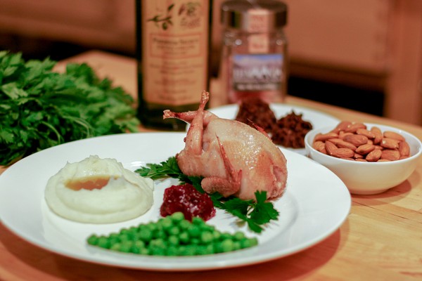 A small cooked bird on a plate along with mashed potatoes and gravy, cranberry sauce, and peas; parsley, a jar of olive oil, a jar of salt, a plate of cookies, and a small white bowl of almonds are in the background