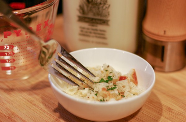 A tiny white bowl filled with some bread crumbs and parsley, being mixed with a fork; a measuring cup, a bottle of milk, and a pepper grinder are in the background
