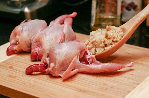 Three small headless, plucked birds on a cutting board; a wooden spoon is being used to shove a mound of stuffing into one of the birds