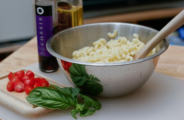 Cooked macaroni in a metal bowl with a wooden spoon in it; basil and tomatoes sit on a wooden cutting board in front of the bowl; vinegar and olive oil are behind the bowl