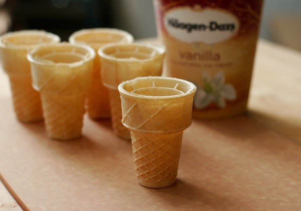 Five flat-bottomed ice cream cones on a table with a pint of vanilla ice cream in the background