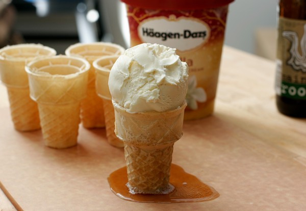 A scoop of vanilla ice cream on an ice cream cone filled with root beer; root beer is leaking out the bottom of the cone and has formed a puddle
