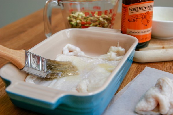 Butter being brushed onto a chicken skin stretched across the bottom of a small, rectangular baking dish next to a pile of chicken skins; a measuring cup of pistachios, and a bottle of orange blossom water are on a cutting board in the background