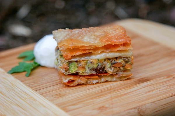 A piece of savoury baklava on a wooden cutting board with a piece of arugula and a small ball of something white behind it