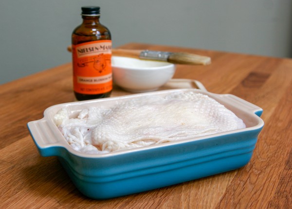 A small, rectangular baking dish full of stretched chicken skins; a bottle of orange blossom water, a brush, and a small white bowl are in the background