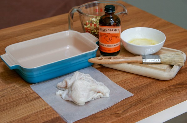 A baking dish, a measuring cup of pistachios, a bottle of orange blossom water, a small white bowl of melted butter, a cutting board, a brush, and a pile of chicken skins on wax paper