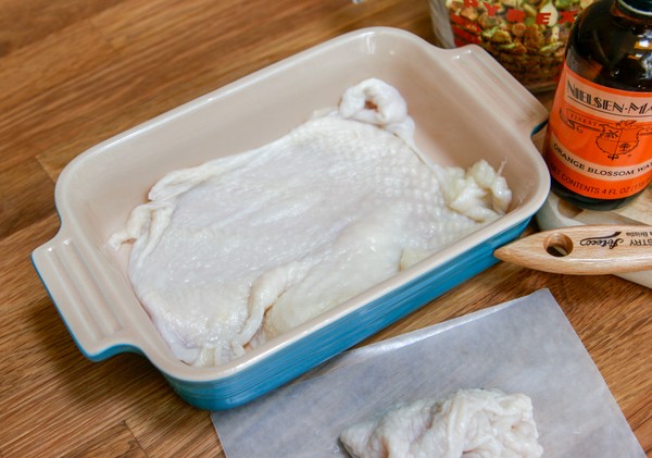 A chicken skin stretched across the bottom of a small, rectangular baking dish next to a pile of chicken skins; a measuring cup of pistachios, a bottle of orange blossom water, and a brush are on a cutting board nearby
