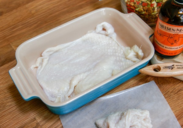 The first layer of chicken skins A chicken skin stretched across the bottom of a small, rectangular baking dish next to a pile of chicken skins; a measuring cup of pistachios, a bottle of orange blossom water, and a brush are on a cutting board nearby