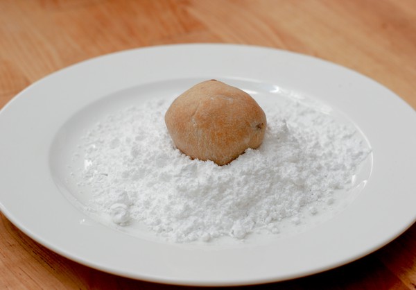 A single, brown, potato-shaped, cookie sitting atop a mound of confectioner's sugar on a white plate