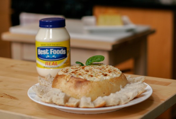 A sourdough bread bowl filled with crab dip and topped with a sprig of basil with a jar of mayonnaise in the background