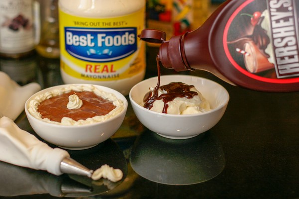 A small white bowl of what appears to be chocolate pudding, next to another small white bowl filled with mayonnaise upon which chocolate syrup is being poured; a jar of mayonnaise is in the background