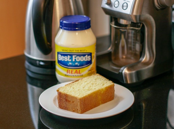 A slice of yellow loaf cake on a white plate with a jar of mayonnaise in the background, along with a coffee grinder and electric kettle